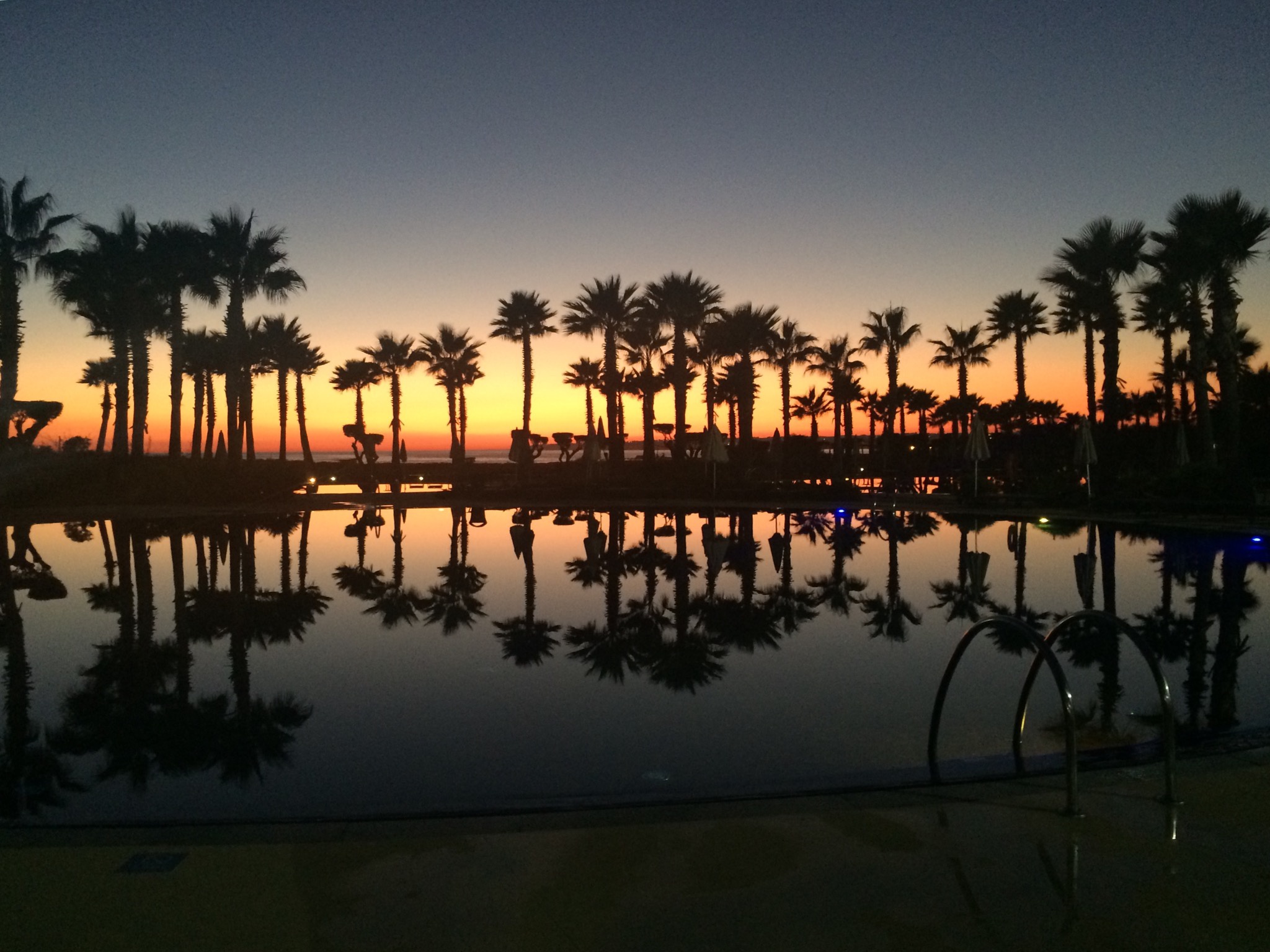 A serene sunset Salgados, Algarve, with the silhouette of palm trees lining the horizon and their reflections in the calm waters of a resort's pool, encapsulating the peaceful ambiance of a luxury escape.